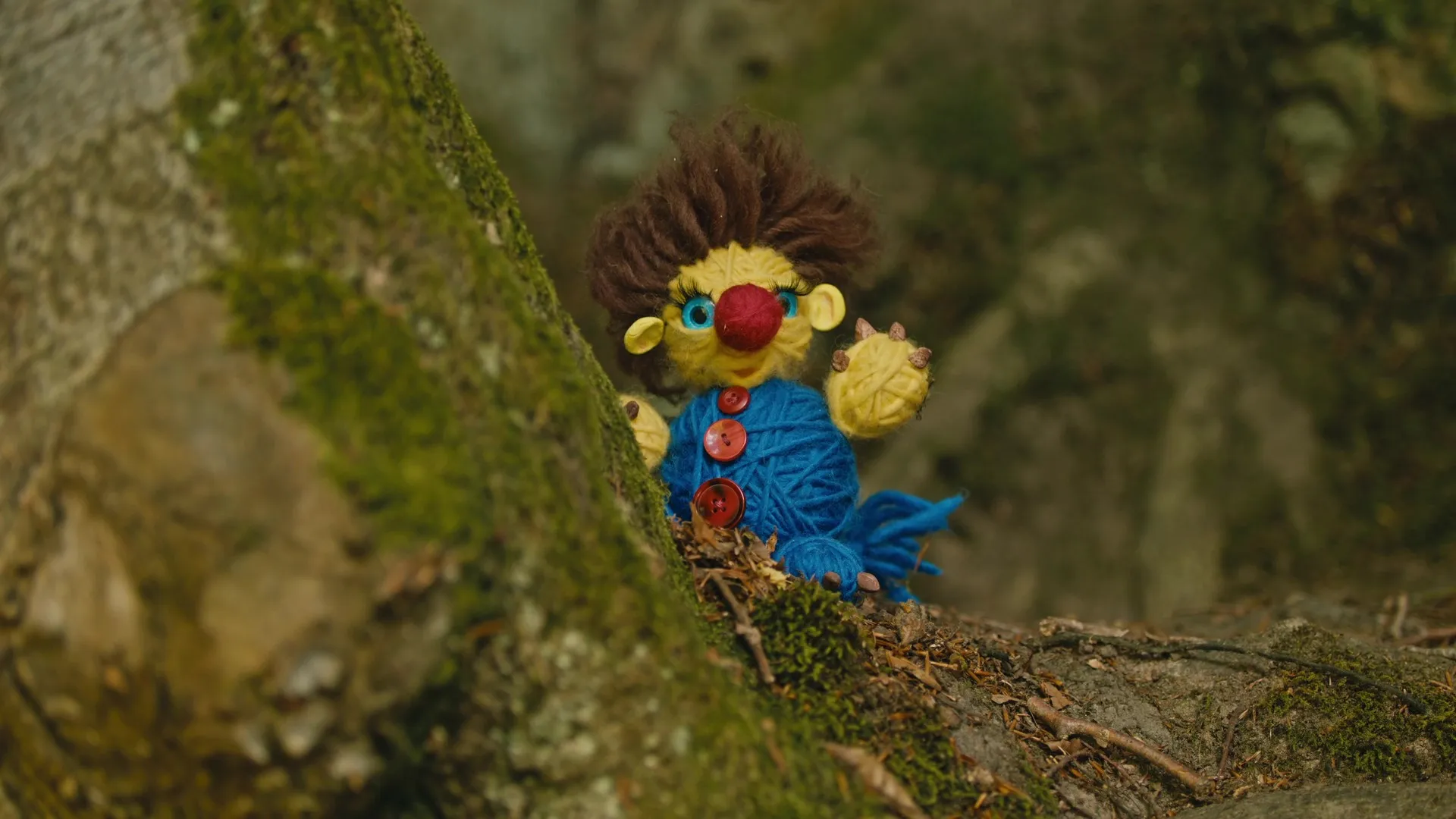 Wolltroll, ein buntes Wollwesen aus Garn und Knöpfen, lugt zwischen moosbedeckten Steinen hervor